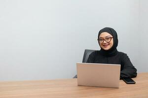 Portrait of attractive Asian hijab woman working on her laptop. Muslim girl doing task in office. Employee and freelance worker concept. photo