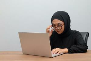 Portrait of attractive Asian hijab woman working on her laptop in office. Muslim girl adjusting glasses to see the screen clearly. Employee and freelance worker concept. photo