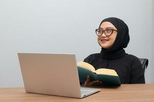 Portrait of attractive Asian hijab woman working on her laptop. Muslim girl reading a book in office. Employee and freelance worker concept. photo