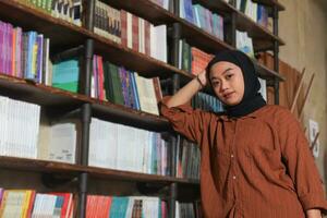 Portrait of Asian hijab woman holding book in front of library bookshelf. Muslim girl reading a book. Concept of literacy and knowledge photo