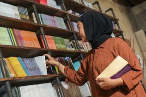 Portrait of Asian hijab woman holding book in front of library bookshelf. Muslim girl reading a book. Concept of literacy and knowledge photo