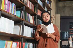 Portrait of Asian hijab woman holding book in front of library bookshelf. Muslim girl reading a book. Concept of literacy and knowledge photo