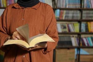 Close up portrait of Asian hijab woman holding book in front of library bookshelf. Muslim girl reading a book. Concept of literacy and knowledge photo