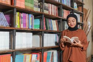 Portrait of Asian hijab woman holding book in front of library bookshelf. Muslim girl reading a book. Concept of literacy and knowledge photo