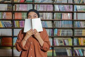 Portrait of Asian hijab woman covering her half head with a book in front of library bookshelf. Muslim girl reading a book. Concept of literacy and knowledge photo