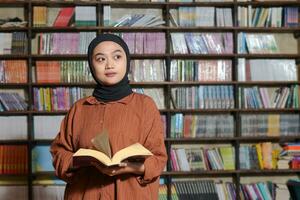 retrato de asiático hijab mujer participación libro en frente de biblioteca estante para libros. musulmán niña leyendo un libro. concepto de literatura y conocimiento foto