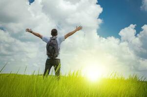 A backpacker on a hill with blue sky and copy space, man backpacking on a green hill with copy space, concept of successful man spreading his hands photo