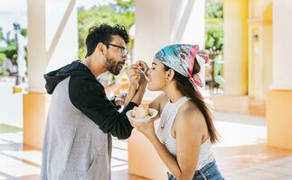 A couple eating shaved ice in a square, Young couple eating shaved ice together with arms intertwined, Side view of young couple with arms linked eating ice cream in a city square photo