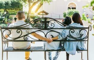 Unfaithful woman sitting holding hands with another man while boyfriend hugs her. Unfaithful couple holding hands on a park bench. Concept of love triangle and infidelity photo