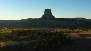 demonios torre butte y carros en la carretera a puesta de sol en otoño. amarillo arboles ladrón condado. Wyoming, EE.UU. aéreo vista. zumbido moscas oblicuo video