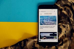KYIV, UKRAINE - 4 MAY, 2023 Zaxid Net ukrainian news portal on smartphone screen with ukrainian flag and camouflage fabric photo