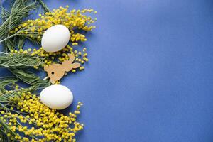 Pascua de Resurrección fondo, blanco huevos y un de madera liebre en un azul fondo, decorado con mimosa flores, plano, parte superior vista, vacío espacio para texto. foto