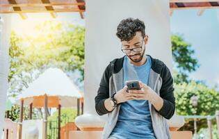 Handsome guy in glasses leaning on a wall chatting on his cell phone, Teenage male leaning on a wall sending a text message, Smiling handsome man using a phone leaning on a wall in a park photo