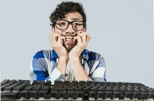 nervioso nerd chico mordiendo su uñas en frente de el teclado, preocupado hombre en lentes a el computadora teclado. joven hombre preocupado cara en frente de teclado. hombre con computadora problemas foto