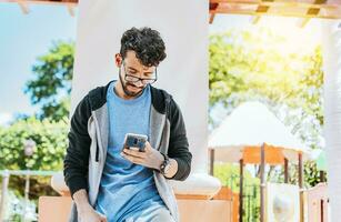 Smiling handsome man using a phone leaning on a wall in a park, Handsome guy in glasses leaning on a wall chatting on his cell phone, Teenage male leaning on a wall sending a text message photo