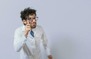 Mad scientist holding a magnifying glass isolated, Man in white coat with magnifying glass looking at camera, Surprised male scientist in lab coat looking at camera with magnifying glass photo