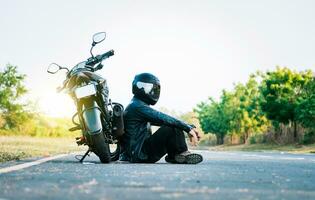 Male biker sitting next to his motorcycle on the road. Male motorcyclist sitting and leaning on his motorcycle on the asphalt photo