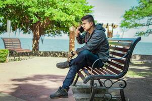 Man sitting on a bench calling on the phone, young man sitting on a bench with cell phone, close up of a young man calling on the phone sitting on a bench photo