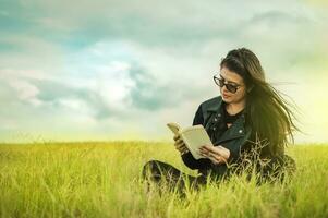 atractivo joven mujer leyendo un libro en el campo, sonriente niña leyendo un libro en el verde campo, adolescente niña sentado en el césped leyendo un libro. concepto de niña en lentes leyendo en el campo foto