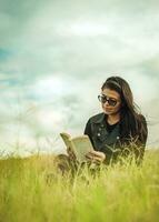 sonriente niña leyendo un libro en el verde campo, adolescente niña sentado en el césped leyendo un libro. concepto de niña en lentes leyendo en el campo, atractivo joven mujer leyendo un libro en el campo foto