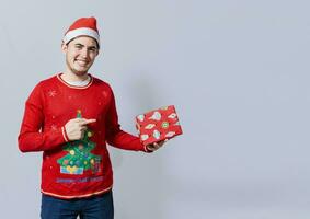 Smiling guy in christmas hat holding and pointing a gift box. Portrait of teenager in christmas hat holding and pointing a gift box. Guy in christmas hat and sweater holding a gift box isolated photo