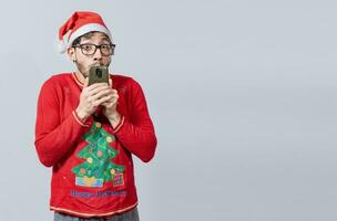 hombre en Navidad sombrero con asombrado cara participación teléfono. en línea Navidad especial ofertas concepto. joven hombre en Navidad ropa y sombrero asombrado participación el teléfono foto