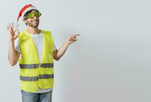 Engineer in christmas hat showing and recommending a product. Builder man in christmas hat pointing a christmas promo. Civil engineer in christmas hat recommending a christmas offer photo