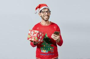 Handsome man in christmas hat holding gift and looking at phone, Guy in christmas hat holding gift box and smiling at smartphone, Christmas man holding gift box and telephone isolated photo