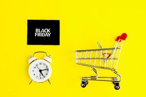 A white alarm clock, a shopping cart, and a black square that says black Friday on a yellow background. The topic of sales. photo