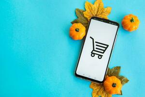 Smartphone with an image of a shopping cart on the screen, pumpkins and maple leaves on a turquoise background. Concept of autumn sales, Cyber Monday, Black Friday. photo