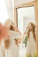 A beautiful teenage girl with long hair measures a straw hat in front of a mirror. Self-admiration of a blonde. photo