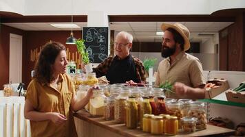 Elderly vendor assisting clients in eco friendly shop with ethical sourced food items, providing recommendations. Woman and man in local store chatting with trader about bulk products video