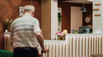 Old people ring service bell at hotel, receptionist ensuring easy check in process and helping tourists with room reservation. Couple on retirement trip arriving at all inclusive resort. Tripod shot. video