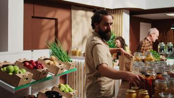 Hipster man in zero waste supermarket using paper bag to carry pantry staples to checkout counter. Healthy living customer in eco friendly local grocery store doing shopping for cooking ingredients video