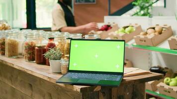 Greenscreen laptop in local eco store with crates of organic homegrown produce and bulk products in jars on shelves. PC running isolated display with mockup copyspace template. video