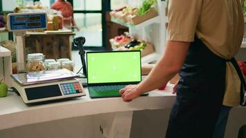 Shop owner works with greenscreen on laptop, sitting at register counter and waiting to serve customers in local zero waste supermarket. Vendor using blank display with chromakey mockup. video