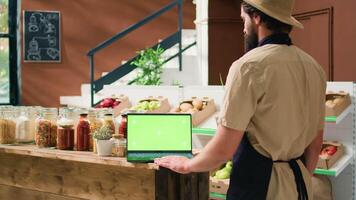 Locale negozio venditore usi schermo verde vicino scaffali pieno con fresco maturo produrre e cibo forniture, guardare a il computer portatile in esecuzione isolato chiave cromatica modello. magazziniere prepara merce per saldi. video