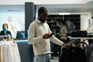 African american man scrolling clothing store online website on smartphone while shopping in department mall. Boutique customer checking shirt size in stock using mobile phone app photo