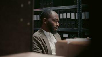 African american man working on criminal case with files, analyzing suspicious evidence and documents in archive space. Young inspector conducting professional investigation. Handheld shot. photo