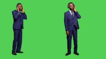 Male manager using headset listening to music on camera, having fun with radio songs over full body green screen. Young businessman in suit dancing and wearing headphones in studio. photo