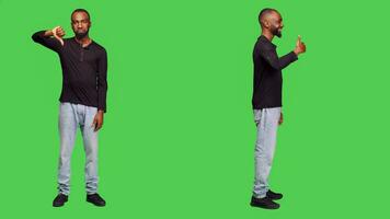 Young adult giving thumbs up and thumbs down in studio, showing approval and disapproval over full body greenscreen. Male person feeling confident doing like and dislike symbol. photo