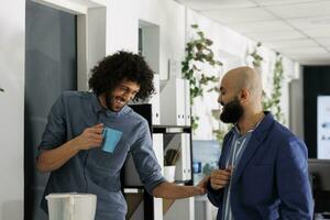 Corporate colleagues telling joke and laughing while drinking coffee in coworking space. Smiling young arab start up company employees enjoying work break in business office photo