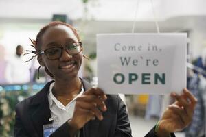 Clothing store smiling african american woman assistant hanging opening signboard on entrance door. Cheerful fashion shop worker putting sign on window noticing about workday beginning photo