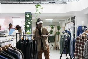 africano americano hombre de influencia grabación promocional vídeo para ropa marca utilizando teléfono inteligente en trípode en Moda boutique. blogger publicidad ropa de caballero en social medios de comunicación para seguidores foto