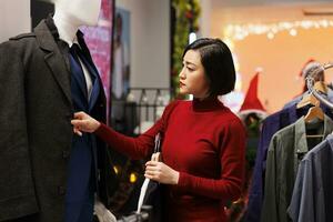 Asian customer browses for clothes on sale, looking for new fashion items in clothing store during christmas seasonal promotions. Woman checking fabric of garments on hangers, buying presents. photo