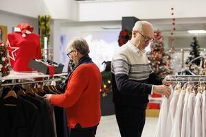 Elderly people searching for clothes in shopping center, looking at fashion items on hangers during winter season. Couple checking new merchandise in retail store, buying presents for family. photo