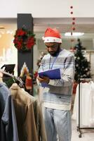 Store employee works on logistics with clipboard of items list, wearing festive hat in shop decorated for christmas season. Retail assistant verifying all merchandise and clothes from hangers. photo