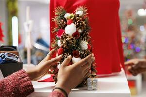 Woman employee arranging small tree festive decor all around clothing store near racks and hangers, personnel decorating christmas ornaments. African american girl putting decorations up. Close up. photo