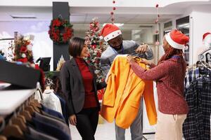 Staff team assisting client in store, showing multiple clothes models to choose from during christmas seasonal sales. African american retail consultants helping asian person to buy perfect items. photo