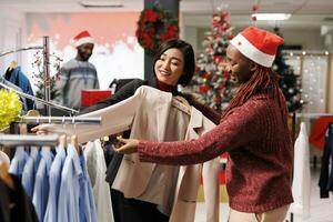 Diverse people looks at items fabric on hangers in retail store at mall, searching for good quality merchandise. Asian woman asking sales assistant about sizes on trendy suit jacket for present. photo